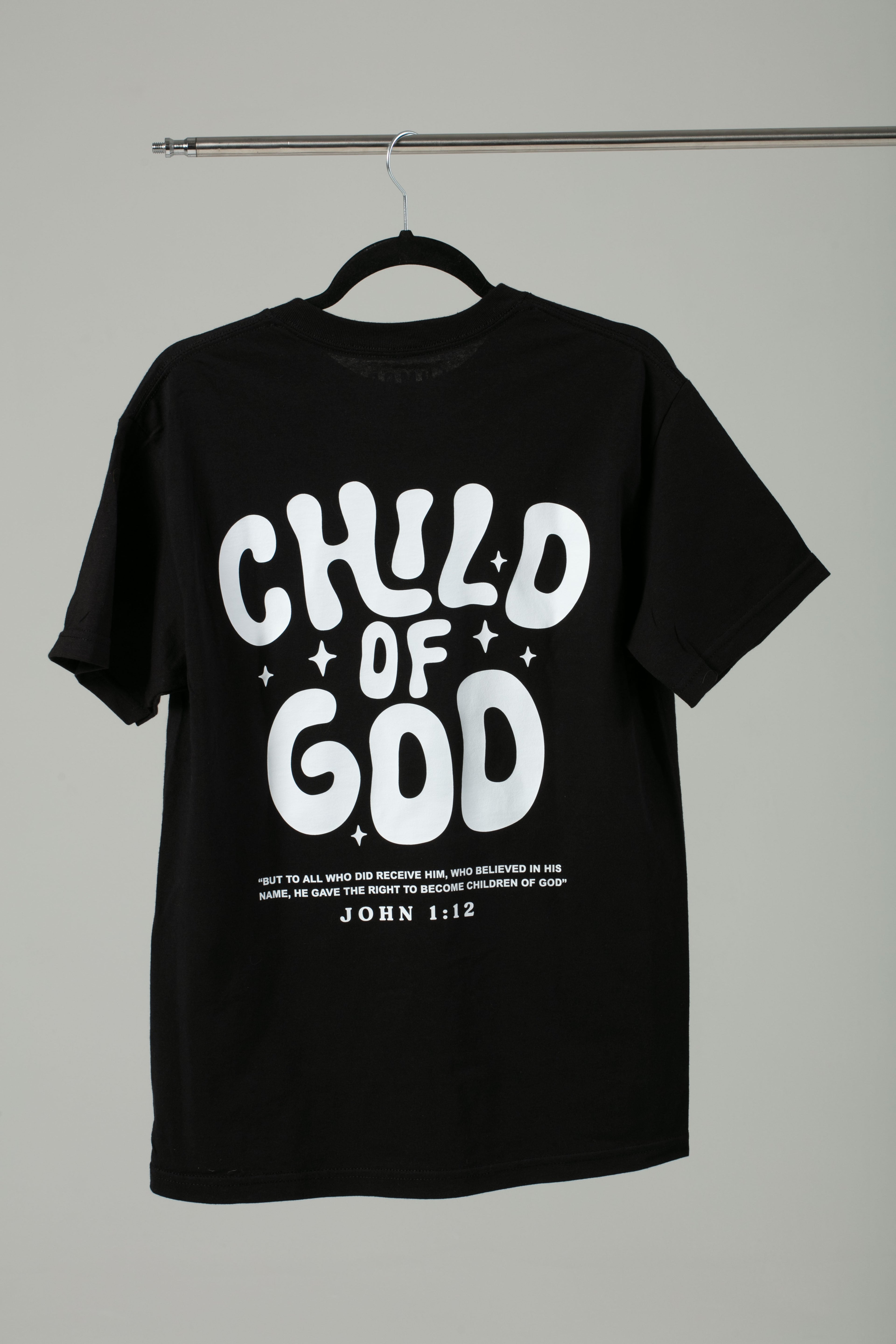 Black ‘Child of God’ t-shirt hanging on a hanger, featuring bold white lettering and John 1:12 on the back.