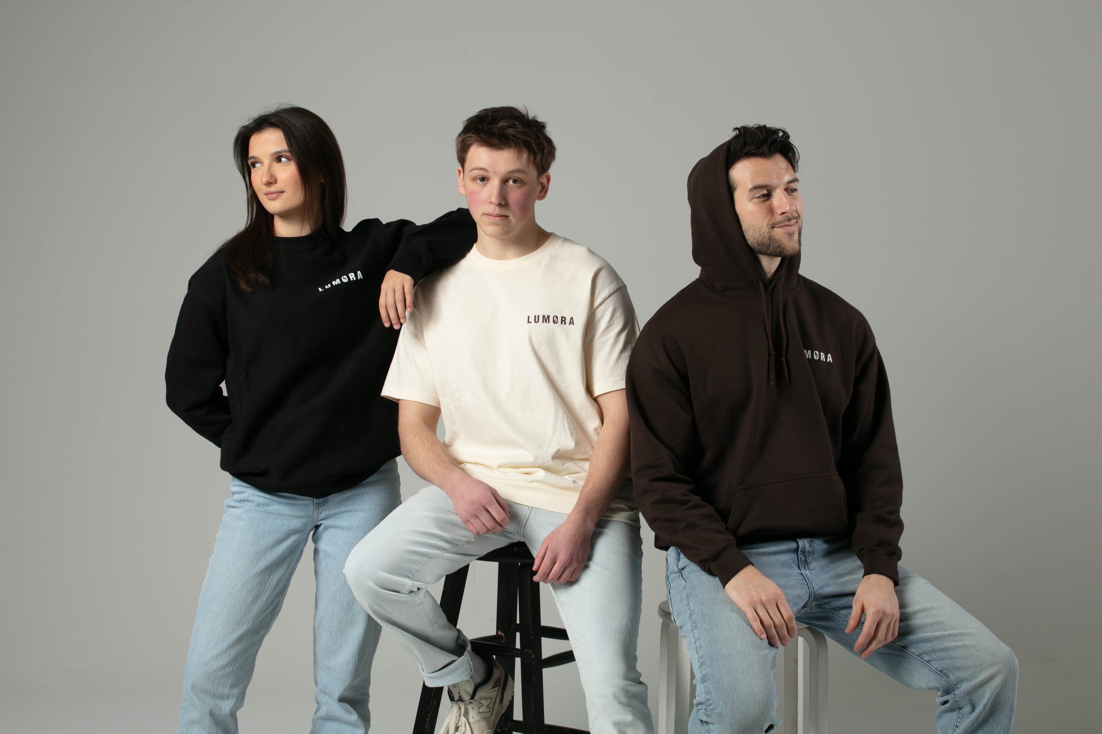 Three models wearing Lumora apparel: black crewneck, cream t-shirt, and brown hoodie, posed together in studio.